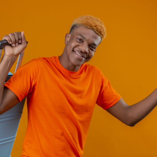 pleased young african american handsome boy wearing orange t-shirt holding travel suitcase and air tickets smiling happy and positive standing over orange background